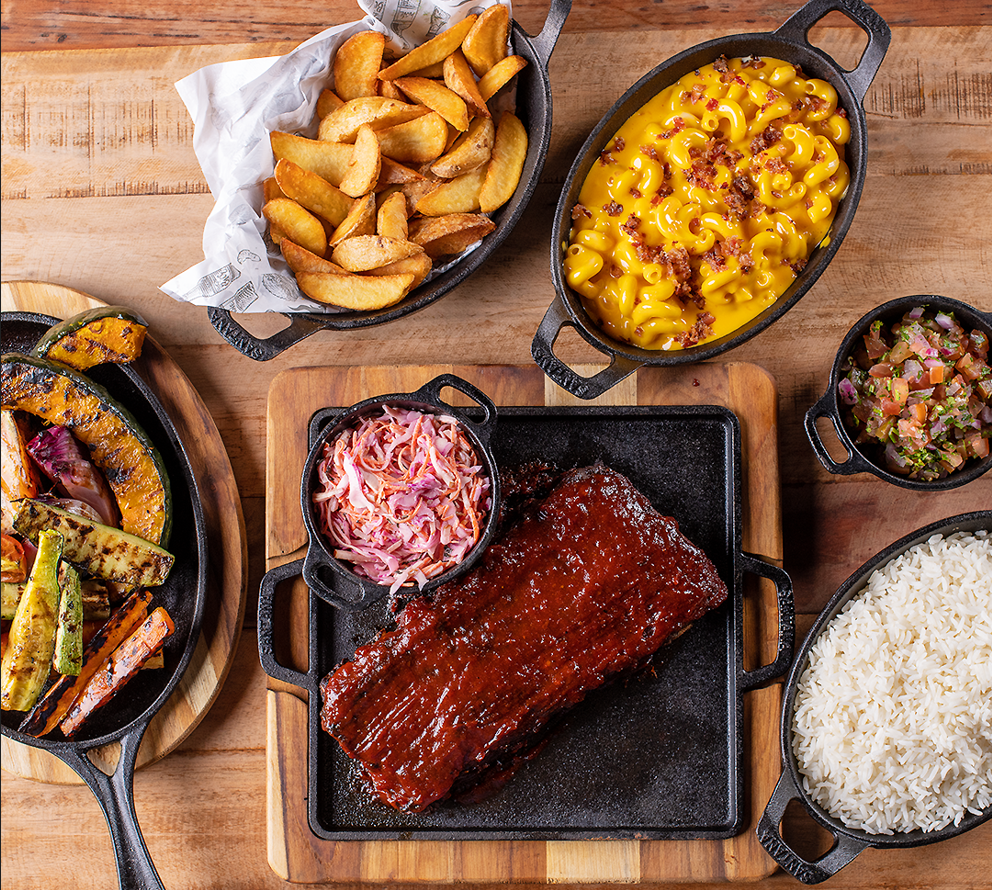 Mesa rústica de madeira com um farto buffet de churrasco para eventos em campinas, destacando uma costela suína ao molho barbecue, mac and cheese, batatas rústicas, legumes grelhados, salada coleslaw, vinagrete e arroz.
