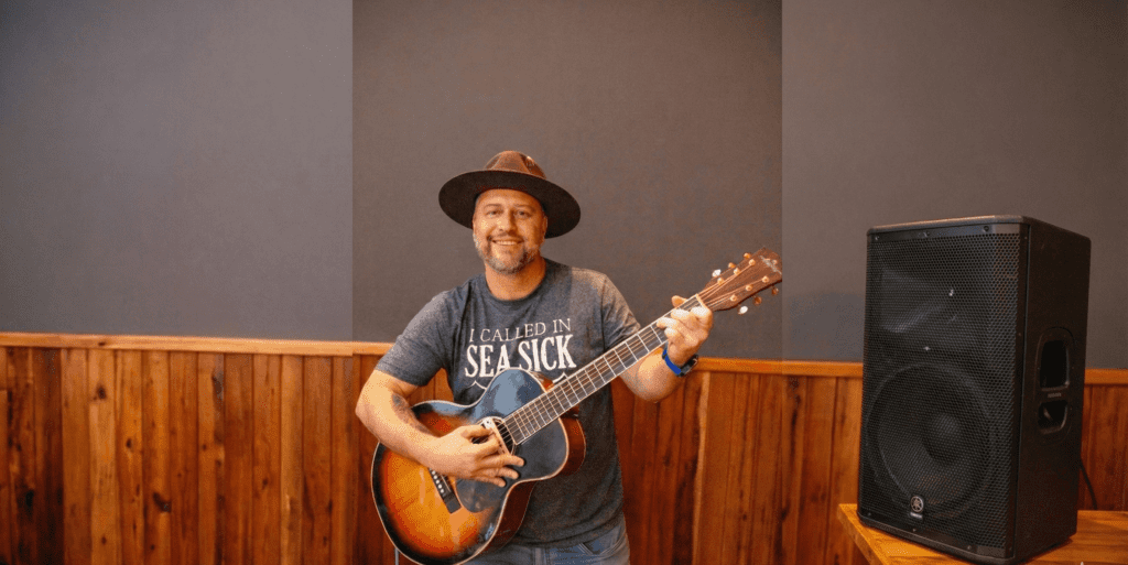 Músico sorrindo com chapéu e violão ao lado de uma grande caixa de som no Ramalho Experience, o melhor restaurante com música ao vivo em campinas para suas noites de sexta e sábado.