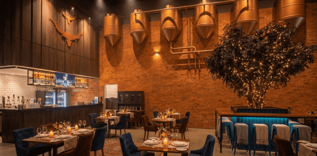 Uma visão ampla e acolhedora do interior de um restaurante romÂntico em campinas, sofisticado com microcervejaria, mostrando mesas de jantar postas com velas e cadeiras de veludo, um bar totalmente abastecido com garrafas de álcool, e uma grande árvore artificial com luzes de fada no centro. Ao fundo, uma fileira de grandes tanques de cobre expostos na parede de tijolos destaca a microcervejaria.