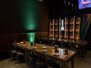 Mesas longas de madeira organizadas com pratos e talheres no salão principal do espaço para eventos em Campinas Ramalho Experience, com iluminação ambiente e barris de chopp ao fundo.