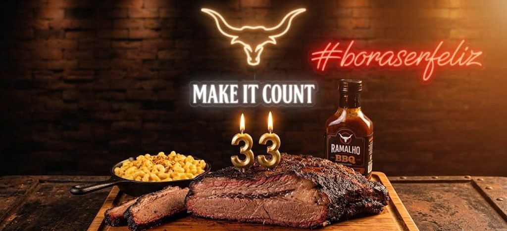 Uma tábua rústica com brisket defumado e velinhas de aniversário acesas, com letreiros de neon ao fundo escrito '#boraserfeliz' e 'MAKE IT COUNT